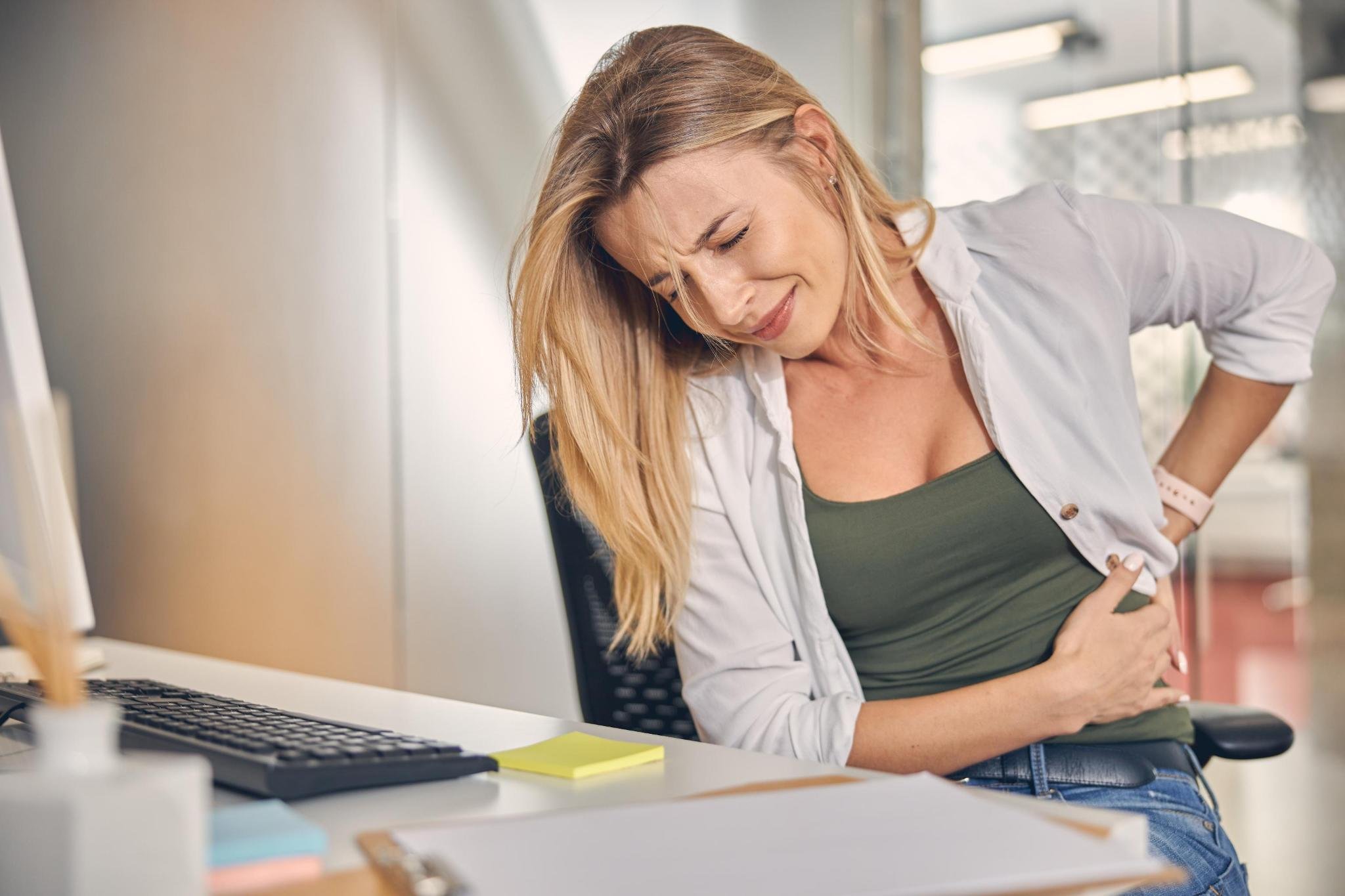 A woman at her office desk wincing and holding her lower back in pain