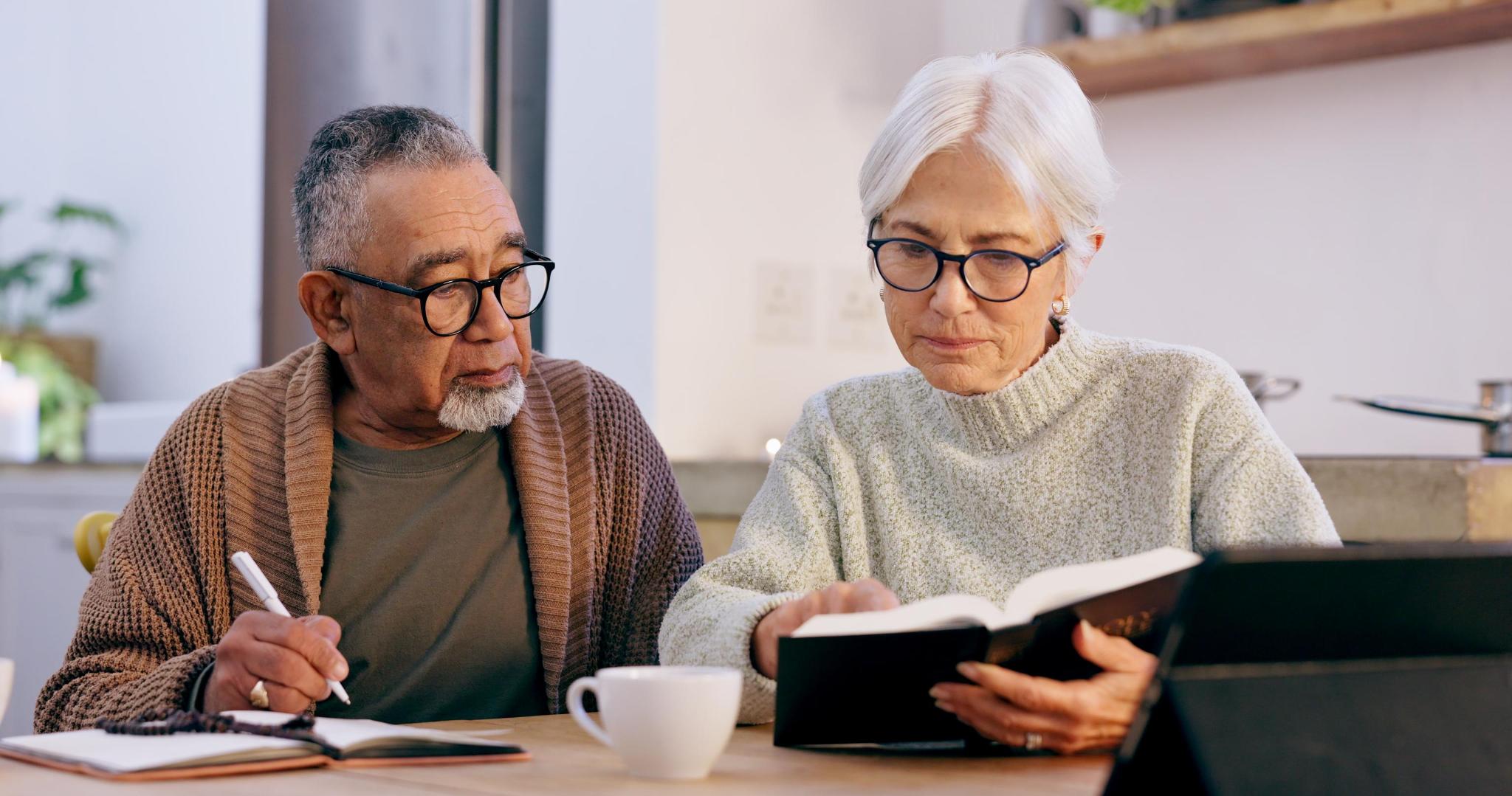 An older couple at their kitchen table reviewing paperwork together, one taking notes and one reading